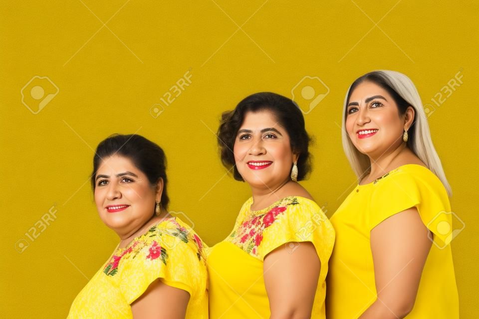 Abuela, hija y nieta con blusas con estampado floral en fila mirando a la cámara, tres generaciones de mujeres mexicanas de fondo amarillo