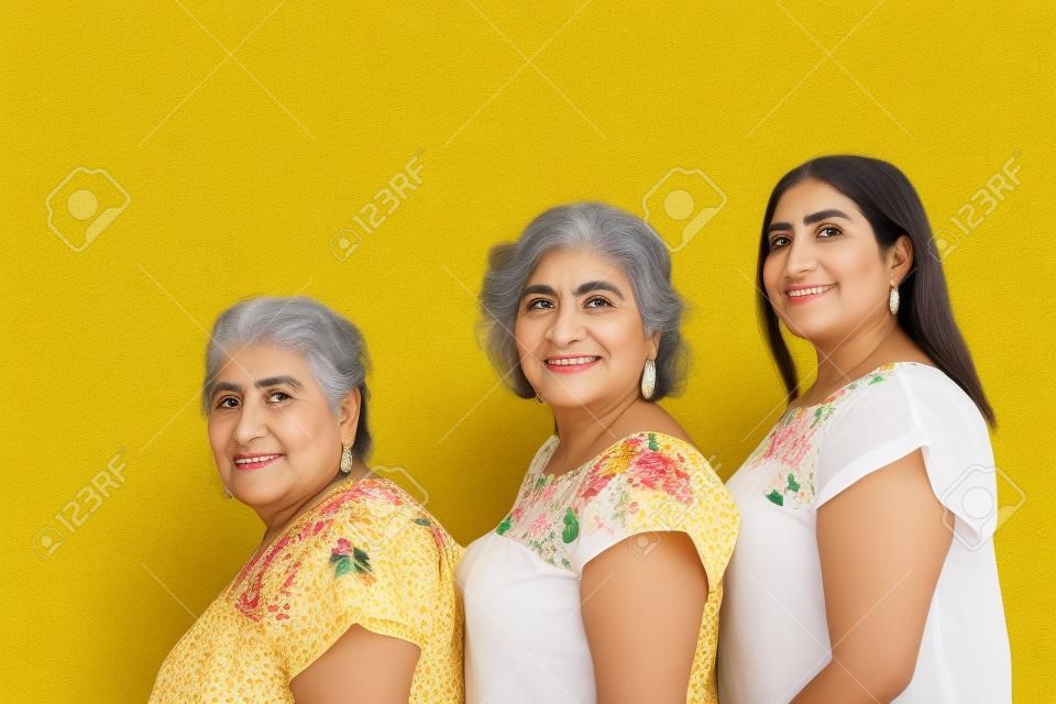 Abuela, hija y nieta con blusas con estampado floral en fila mirando a la cámara, tres generaciones de mujeres mexicanas de fondo amarillo
