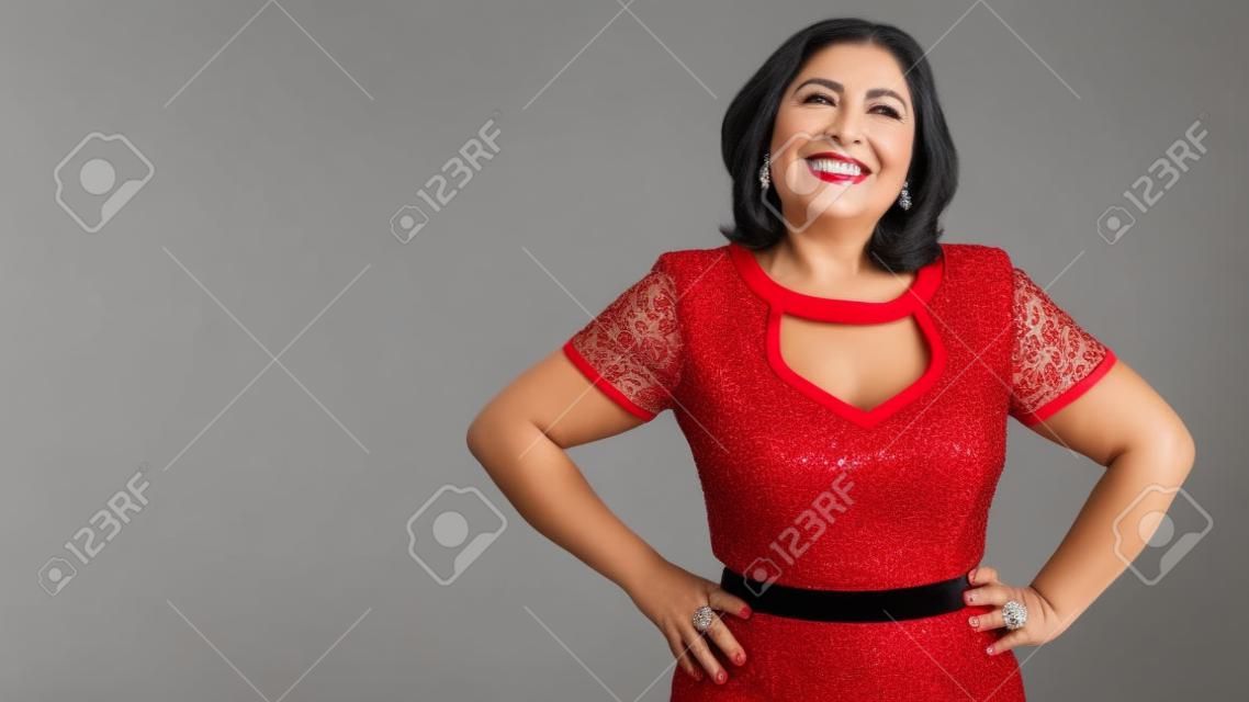 Elegante mujer mexicana madura sonriendo y mirando a la cámara, cabello negro grisáceo, con un vestido rojo con detalles blancos sobre fondo blanco de estudio. Mujer de negocios exitosa. Espacio para texto