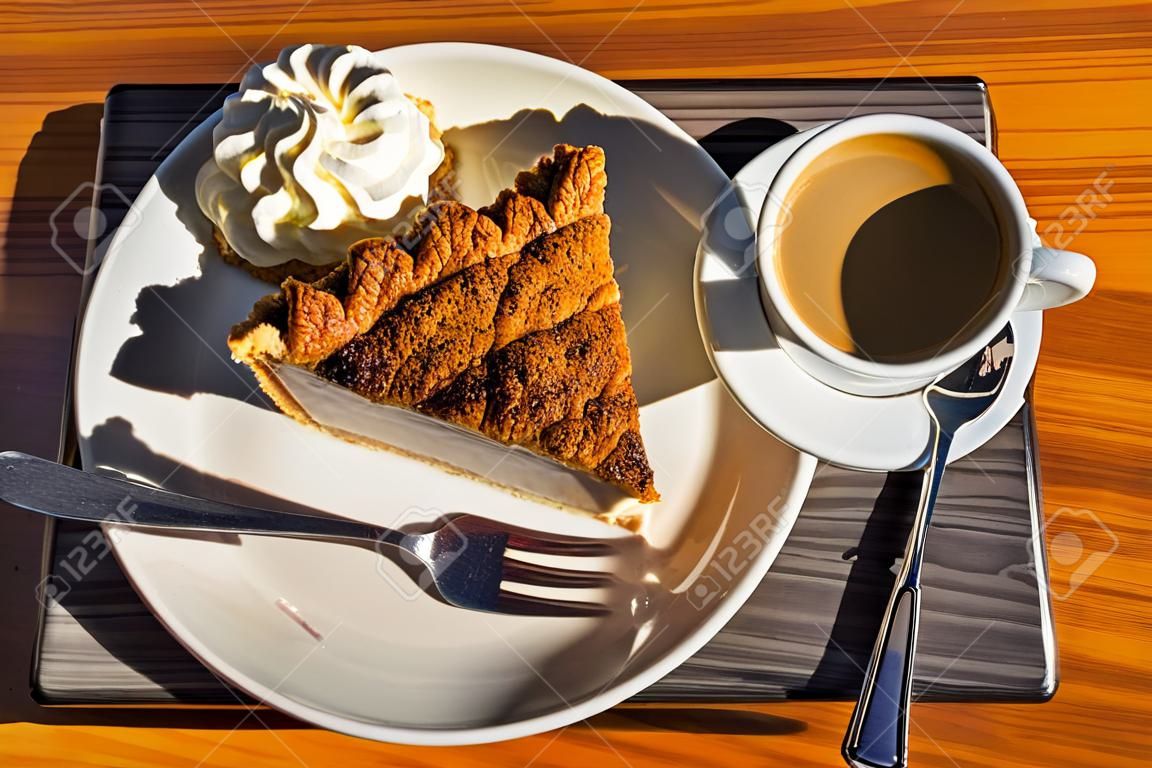Sun shining on and casting shadows of a square white plate with a cup of black coffee and a piece of apple pie with whipped cream