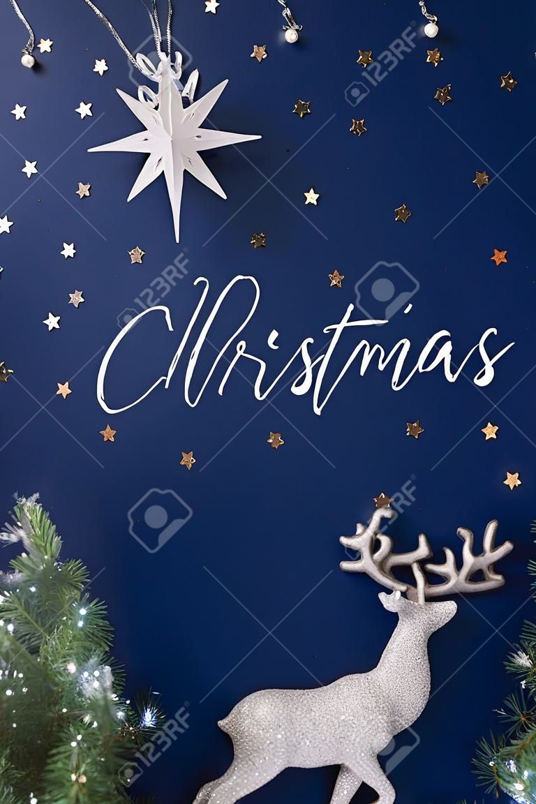 Biglietto di auguri per le vacanze con scritta Merry Christmas. Scena natalizia composta da statuine in argento di renne, stelle e rami sempreverdi su sfondo blu scuro. Concetto di notte di Natale.