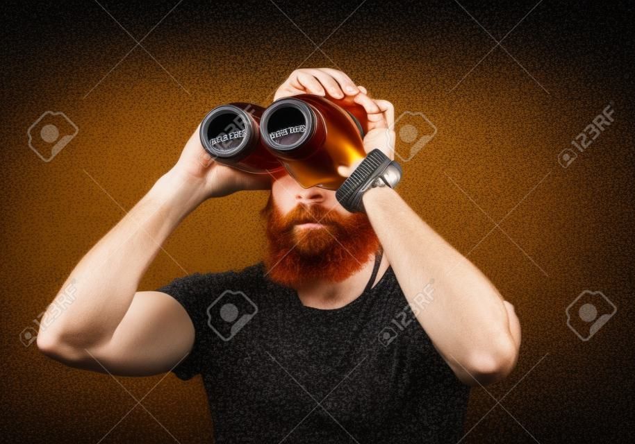 Śmieszne rude Brodaty mężczyzna patrząc przez dwa butelki piwa craft przez lornetkę.