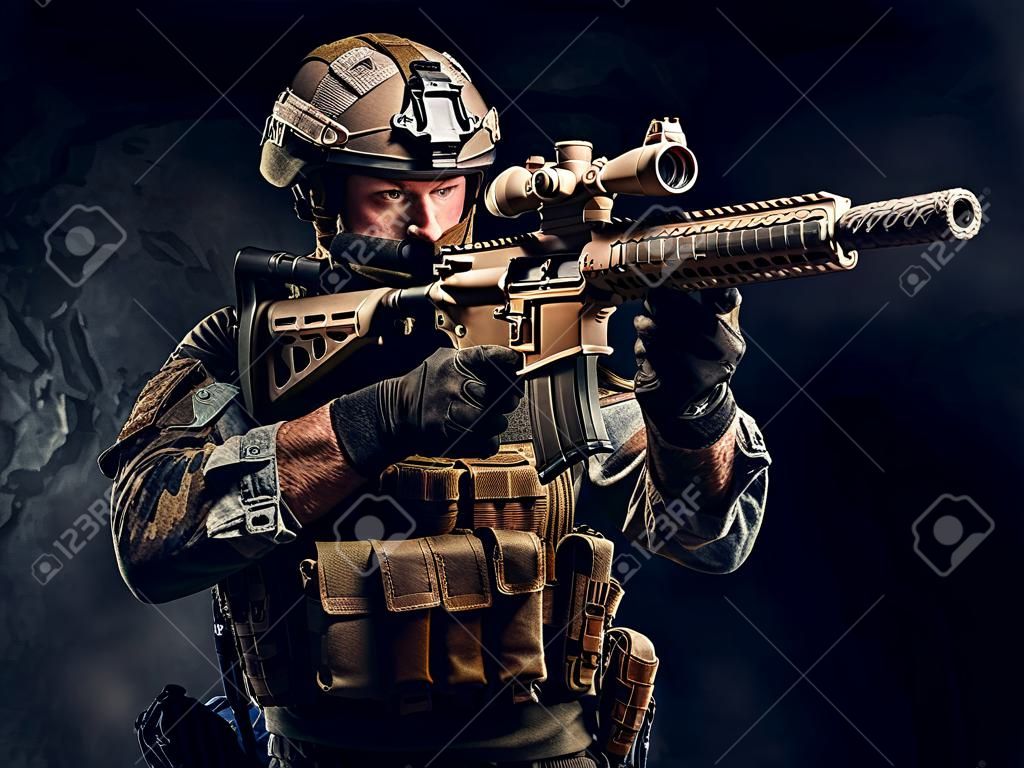 Unidad de élite, soldado de las fuerzas especiales en uniforme de camuflaje sosteniendo un rifle de asalto y apuntando con una mira óptica. Foto de estudio contra una pared con textura oscura