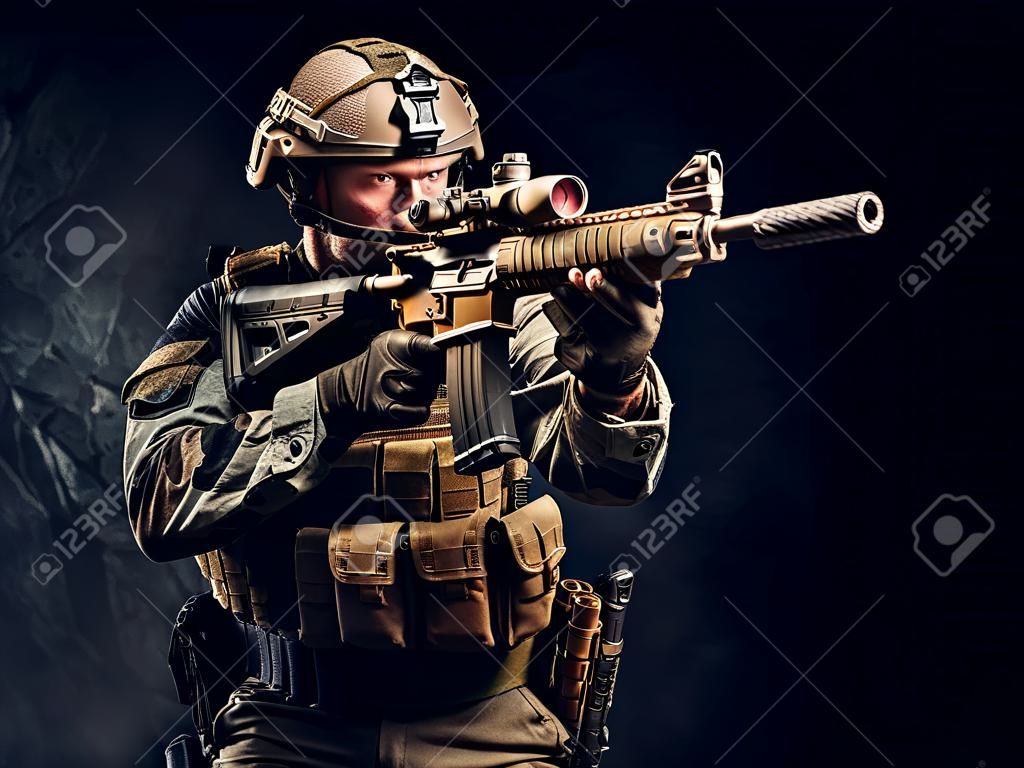 Elite unit, special forces soldier in camouflage uniform holding assault rifle and aiming with optical sight. Studio photo against a dark textured wall