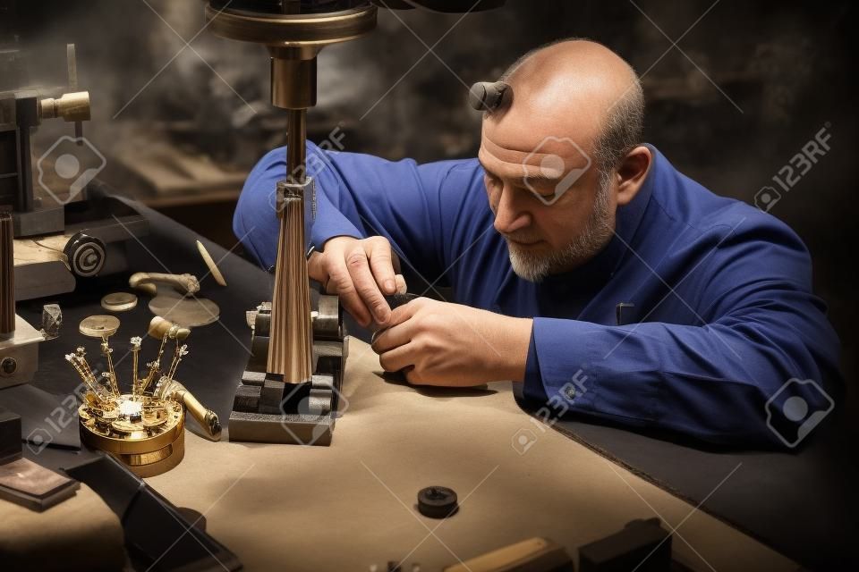 Mature watchmaker is doing engraving for custmers watch at his workshop.