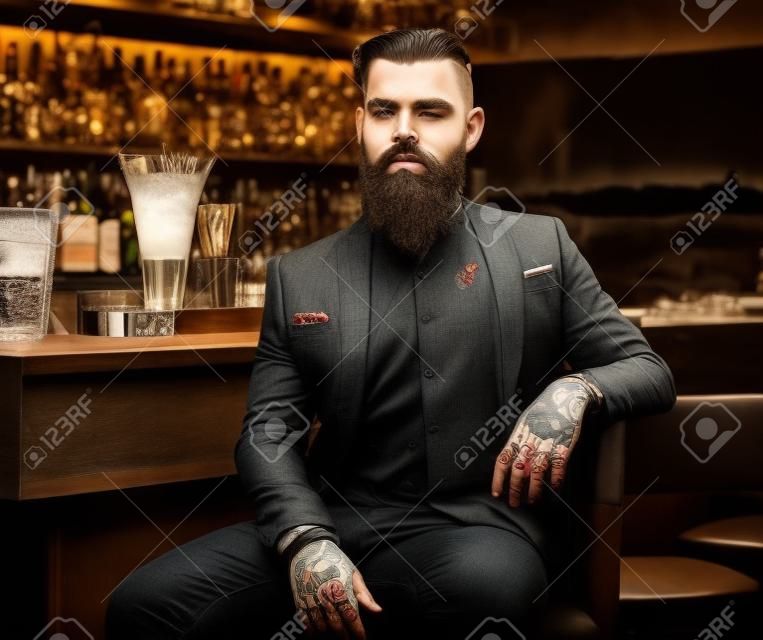 Attraente uomo brutale è seduto vicino al bancone del bar. Ha tatuaggi e barba.