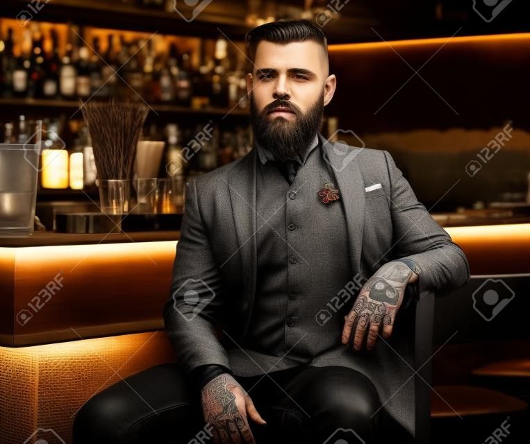 Attraente uomo brutale è seduto vicino al bancone del bar. Ha tatuaggi e barba.