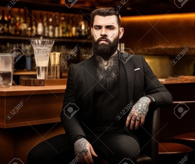 Attractive brutal man is sitting near bar counter. He has tattoes and beard.