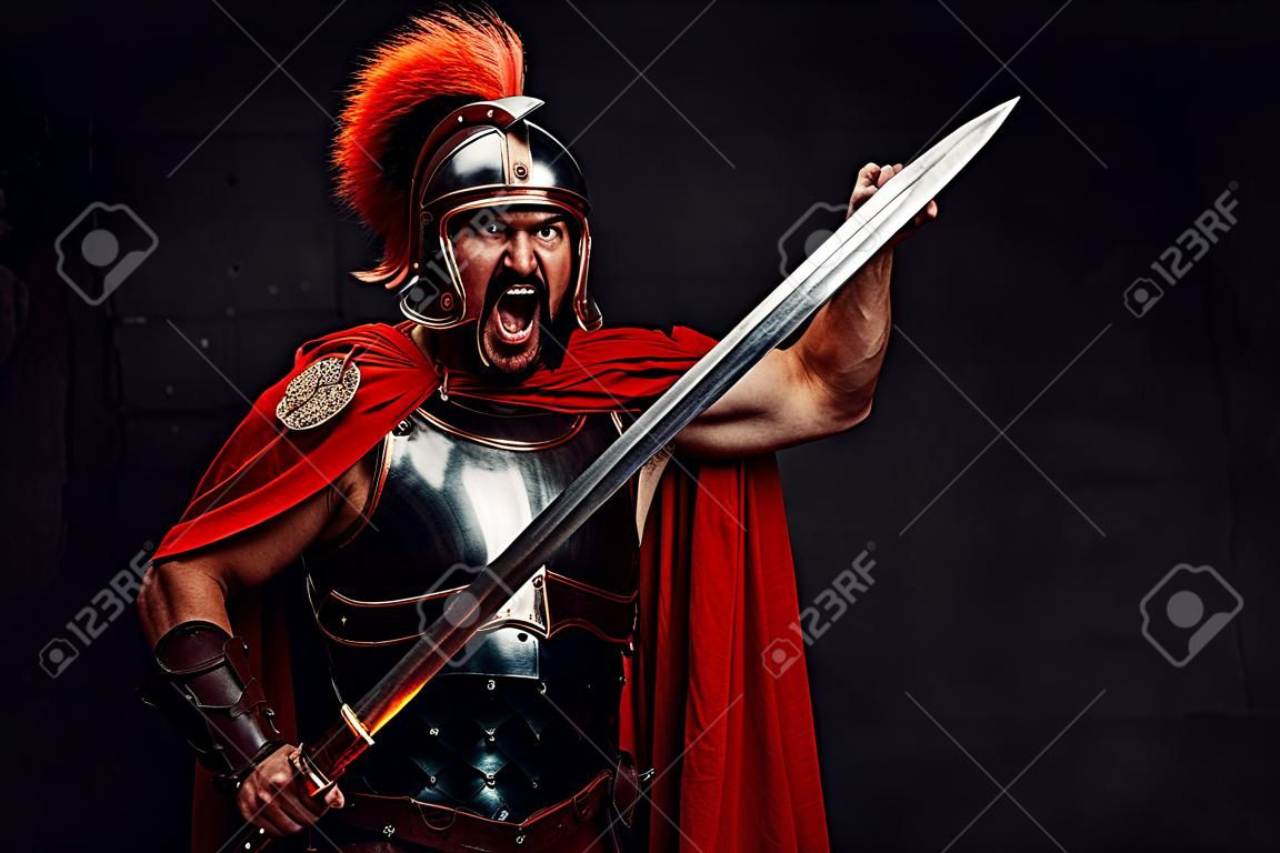 Attacking savage and brutal imperial soldier from rome in steel armour and red cloak holding sword and shield in dark background.