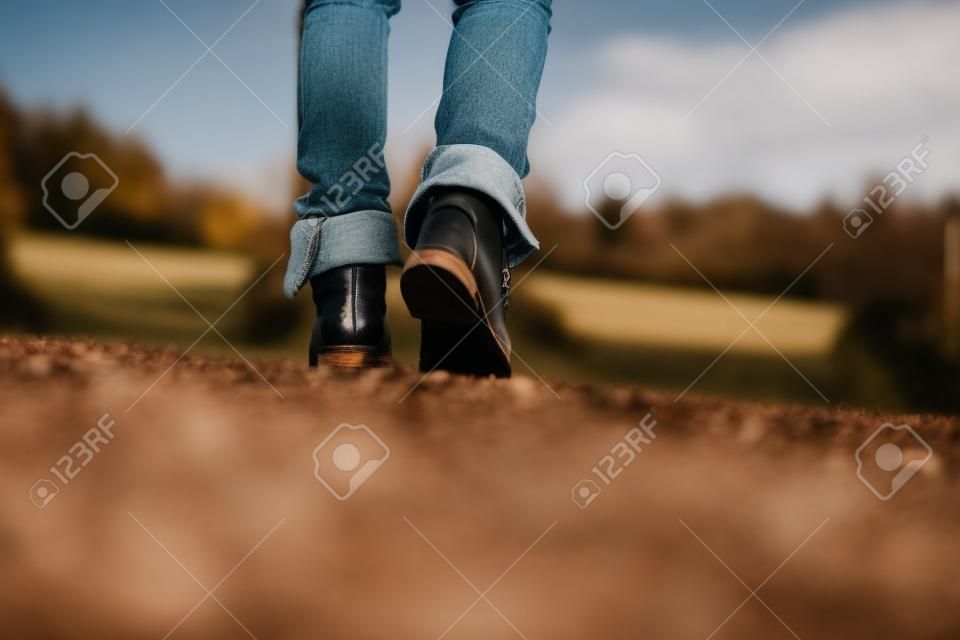 Ángulo de visión de bajo nivel del suelo con el dof bajo de los pies de una mujer con pantalones vaqueros y botas de cuero de alta del tobillo caminando por un sendero rural lejos de la cámara.