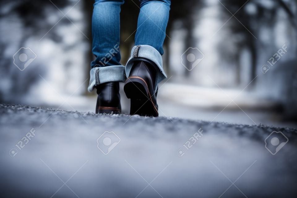 Low angle ground level view with shallow dof of the feet of a woman in jeans and ankle high leather boots walking along a rural path away from the camera.
