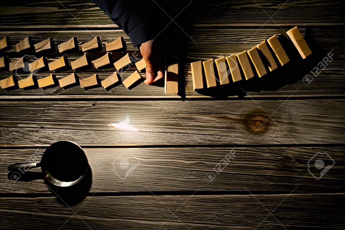 Overhead view of a rustic wooden desk illuminated with a lamp with a seated man stopping the domino effect of falling dominoes by inserting a hand in their path in a conceptual image with copy space.
