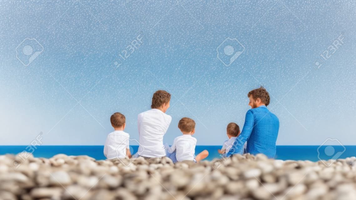 Jonge familie van vijf zittend op een kiezelstrand in het prachtige Griekenland met hun rug naar de camera genietend van hun vakantie.