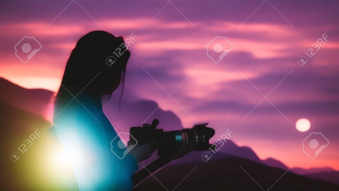 彼女は日の出の光によって照らされた美しい自然の中に立って、プロパティを調整する彼女の一人一人座のカメラを見て若い女性写真家。