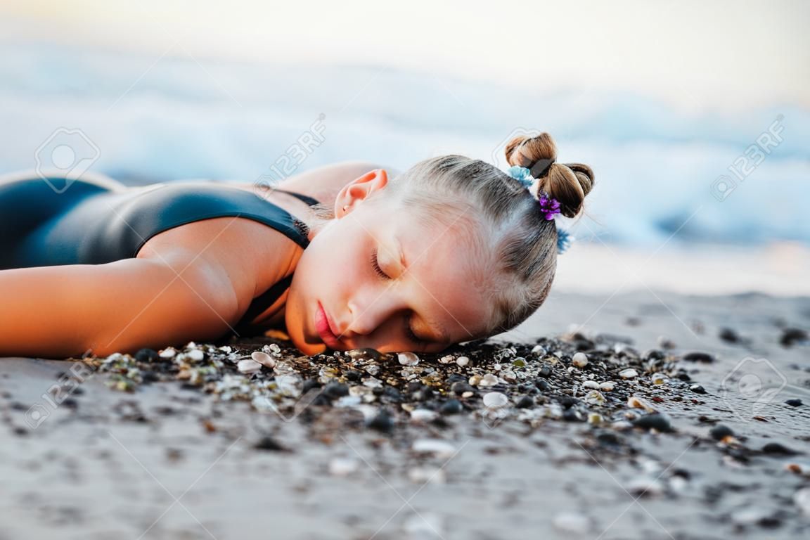 Девушка-подросток сидит на пляже в черном купальнике.