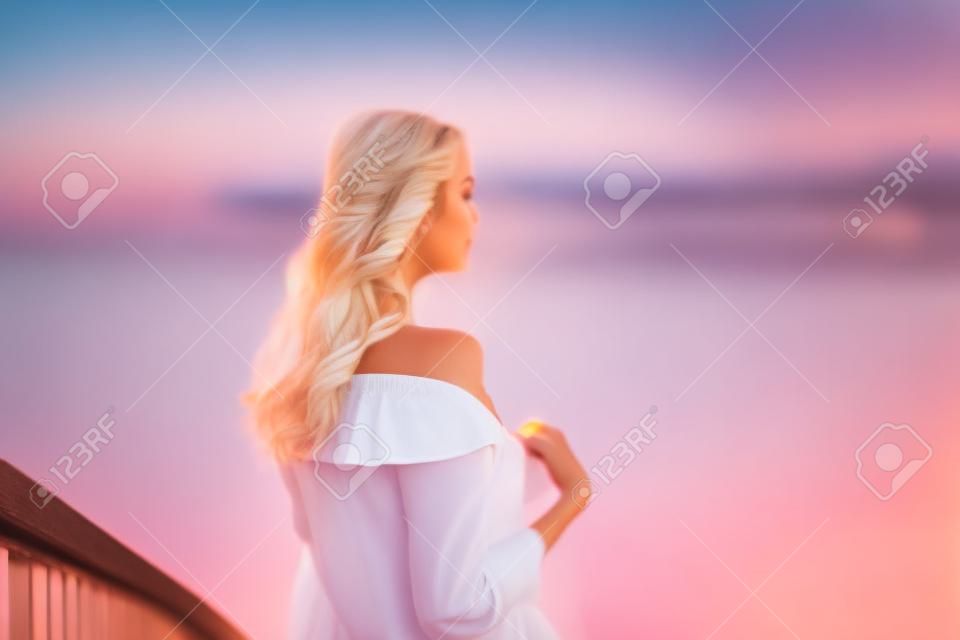 Hermosa joven rubia con camisa blanca en el muelle al atardecer. foto borrosa, fondo