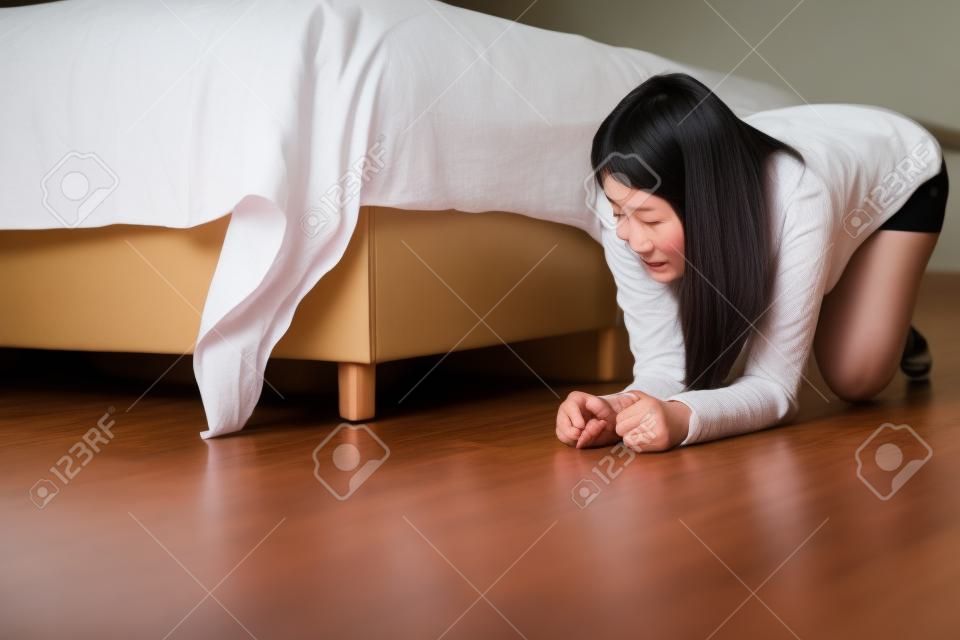 Mujer asiática doblada y buscando algo debajo de la cama, cosa perdida en el dormitorio