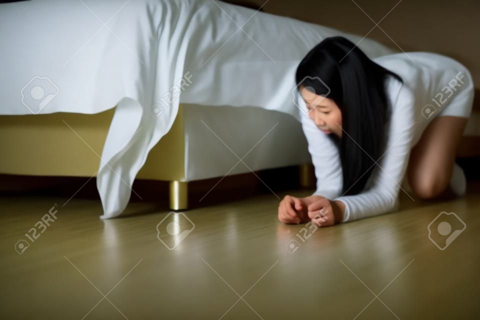 Mujer asiática doblada y buscando algo debajo de la cama, cosa perdida en el dormitorio