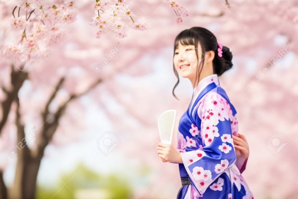 donna in yukata (abito kimono) con in mano un ventaglio pieghevole e guardando il fiore di sakura o il fiore di ciliegio che fiorisce nel giardino