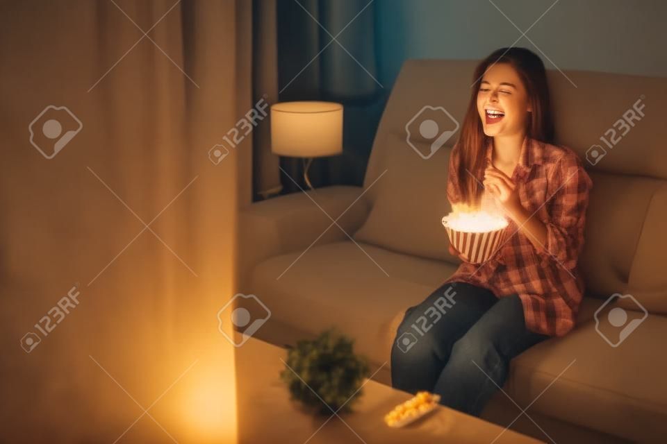 Zabawna młoda kobieta oglądająca telewizję i jedząca popcorn na kanapie w nocy
