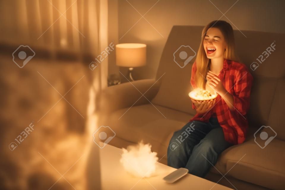 Zabawna młoda kobieta oglądająca telewizję i jedząca popcorn na kanapie w nocy