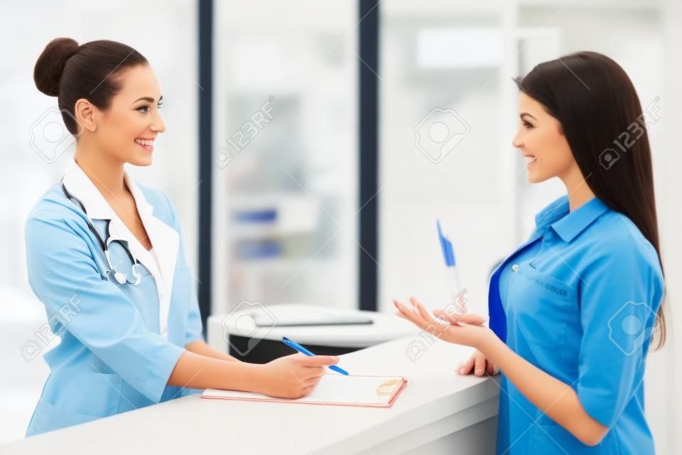 Linda jovem médico feminino está falando com paciente e sorrindo, de pé na sala de espera da clínica