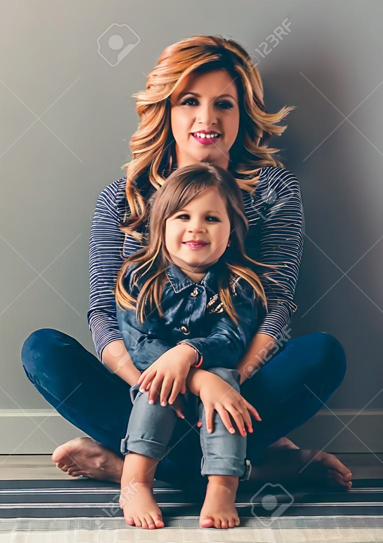 Cute little girl and her beautiful young mom are sitting together on the floor, looking at camera and smiling, on gray background