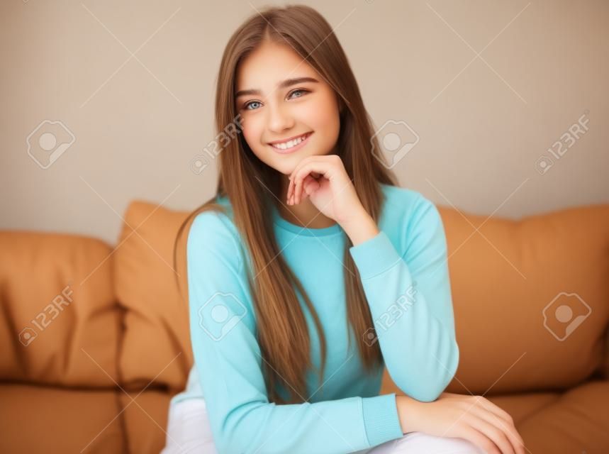 Attractive teenage girls in casual clothes is looking at camera and smiling while sitting on couch at home