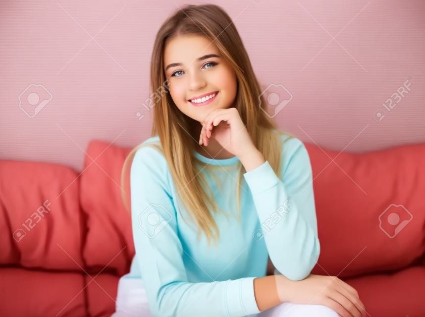 집에서 소파에 앉아있는 동안 캐주얼 의류에 매력적인 십대 소녀는 카메라를보고 웃고 있습니다