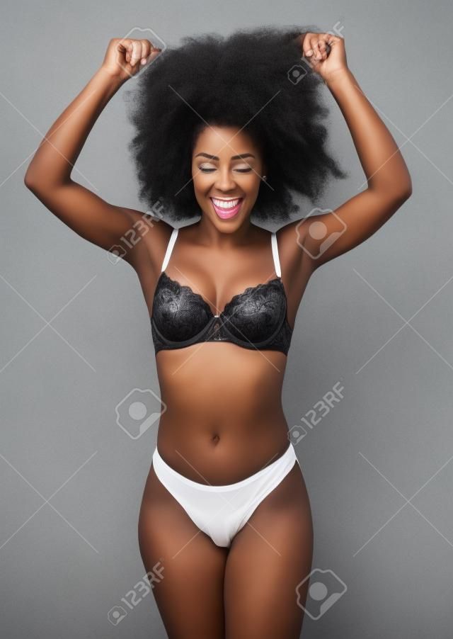 Afroamerykanka w białej bieliźnie bawi się włosami i uśmiecha się, na szarym tle