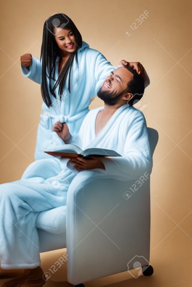 Young man wearing bathrobe sitting in a chair reading book with a woman in robe grabbing his hair with arm pulled back in a fist as if going to punch him