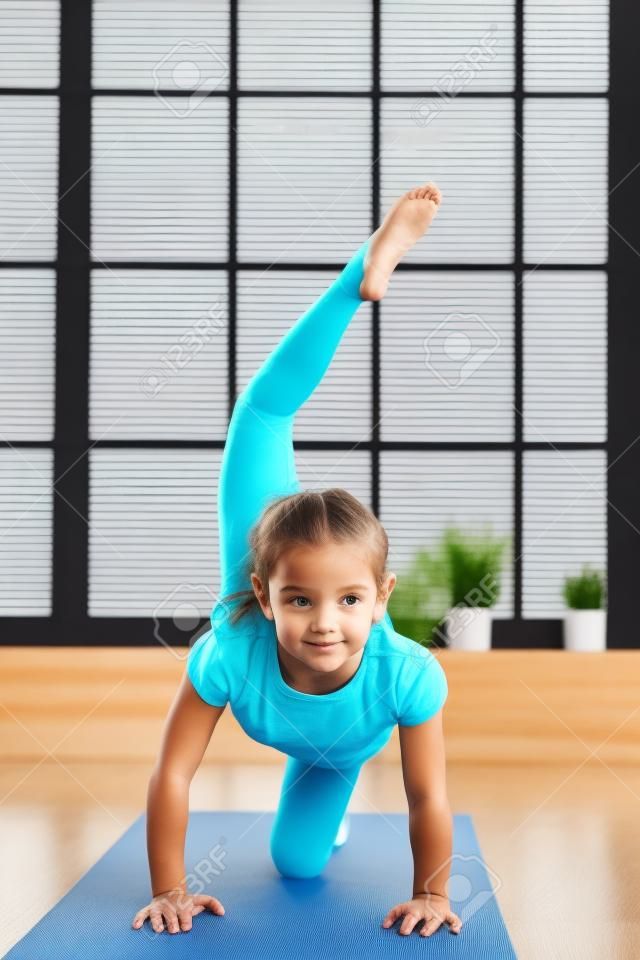 Little girl doing yoga exercise in fitness studio with big windows on background