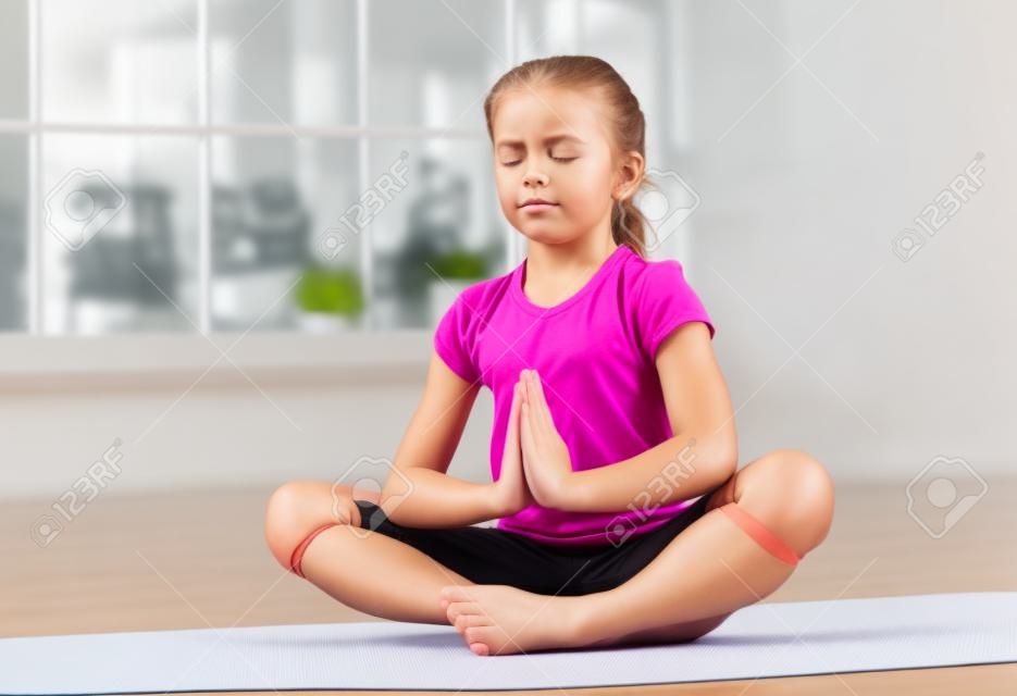 Petite fille assise en position du lotus, les yeux fermés et les mains en studio de remise en forme avec de grandes fenêtres sur le fond