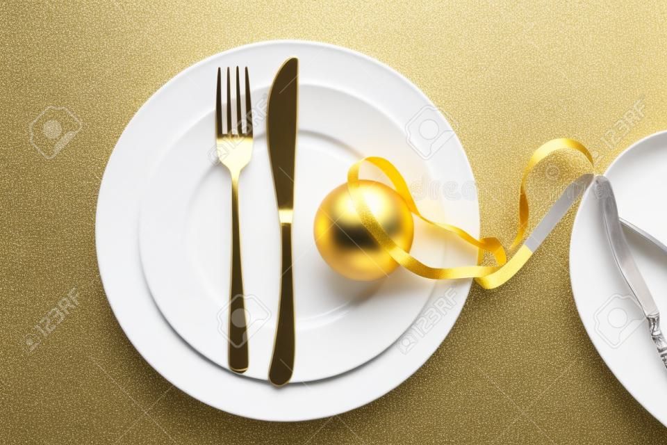 Impostazione della tavola per la cena di Capodanno, palla di Natale d'oro con nastro e posate su set di piatti bianchi, sfondo bianco, vista dall'alto