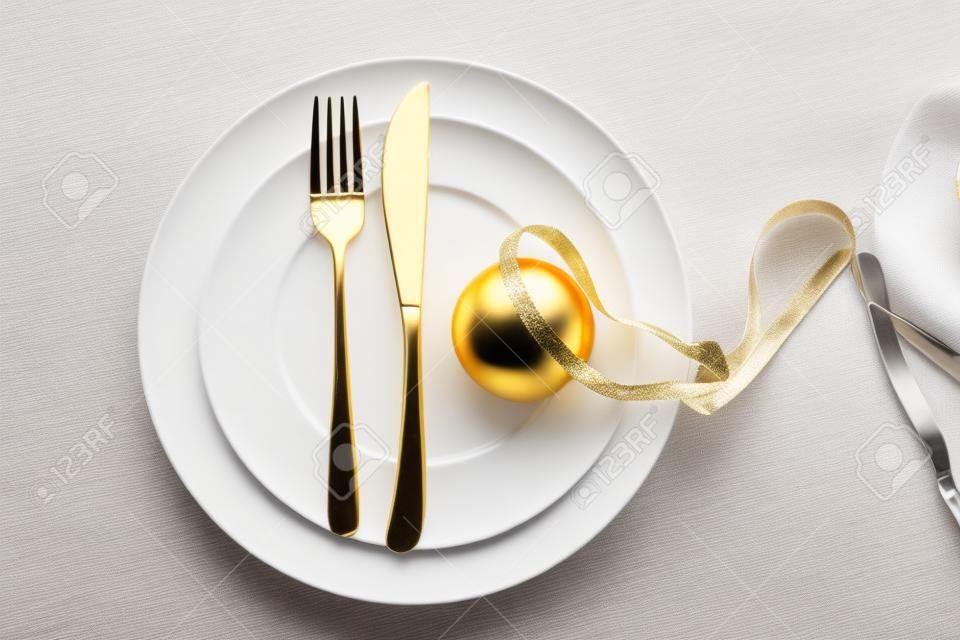 Impostazione della tavola per la cena di Capodanno, palla di Natale d'oro con nastro e posate su set di piatti bianchi, sfondo bianco, vista dall'alto
