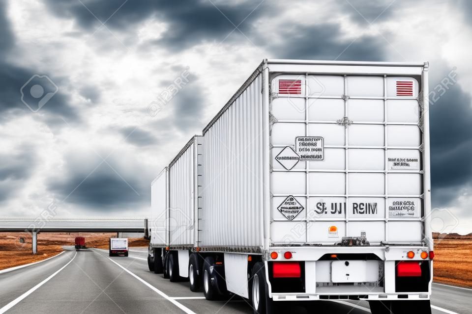 Three trailer road train transporting commercial cargo in USA highway. White long heavy vehicle rear, travels with cloudy sky background.