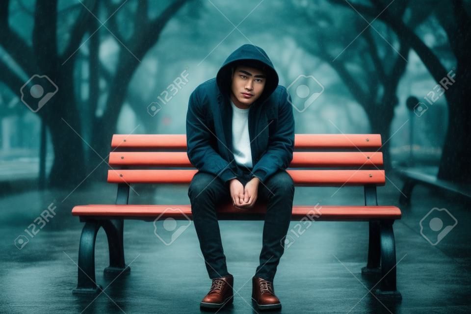 Jeune bel homme hipster asiatique assis sur un banc sous la pluie