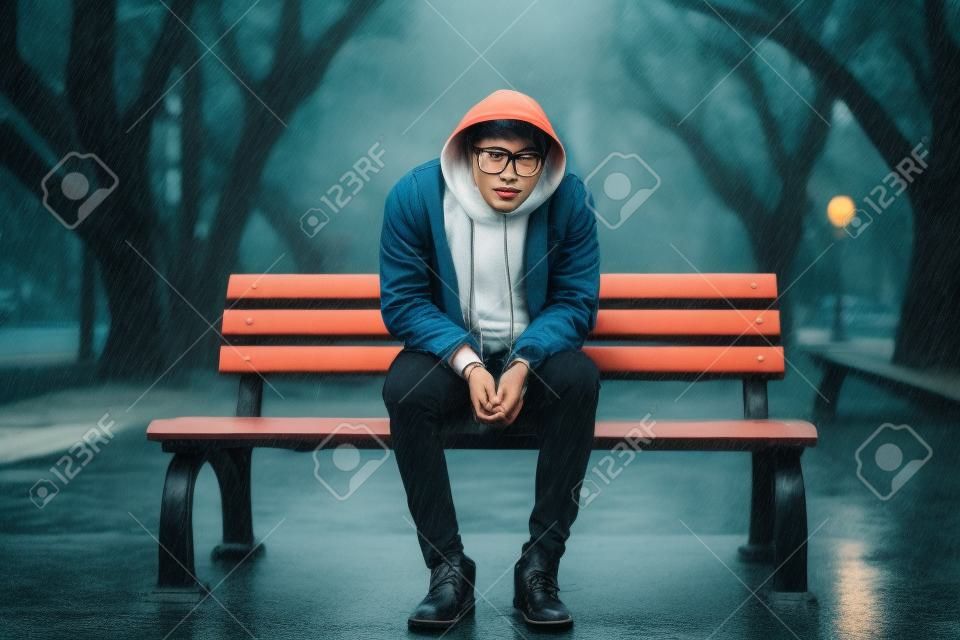 Jeune bel homme hipster asiatique assis sur un banc sous la pluie