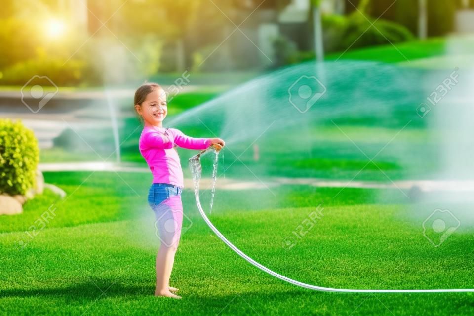 Cute little blong girl watering green lawn. Child pours water from hose on grass at garden. Kid having fun at backyard on bright sunny day. Children summer leisure activity.