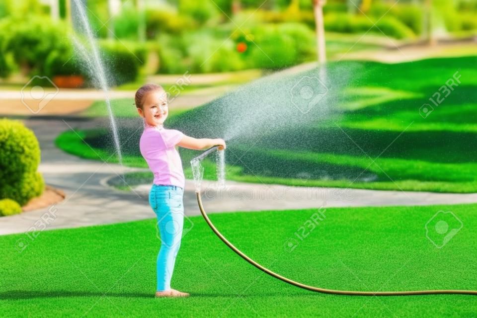 Cute little blong girl watering green lawn. Child pours water from hose on grass at garden. Kid having fun at backyard on bright sunny day. Children summer leisure activity.