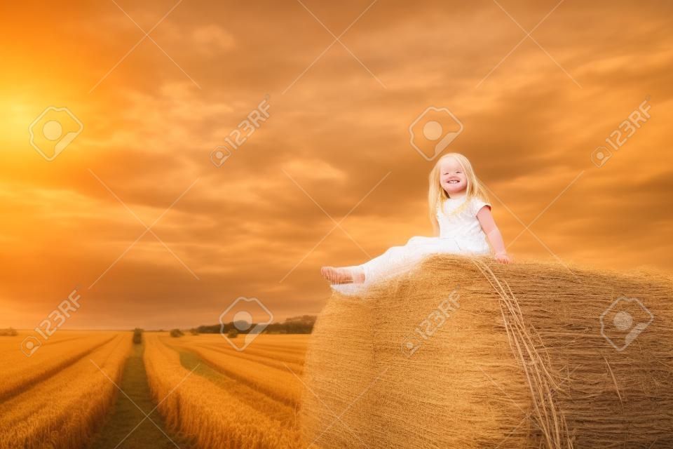 Portret ślicznej małej blond pięknej uroczej wesołej kaukaskiej dziewczynki lubi siedzieć na stosie siana lub beli na zebranym polu pszenicy ciepły letni wieczór. malownicze wiejskie tło krajobrazu kraju