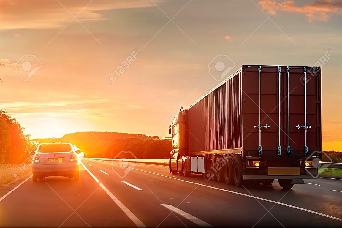 Scenic front view big long heavy semi-treailer truck with sea shipping container driving highway dramatic warm morning evening sunrise sun sky sunset. Cargo transport industry background concept