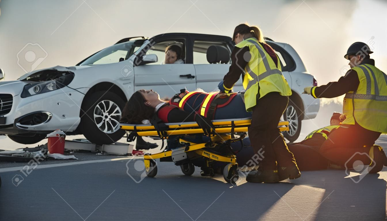 An der Unfallstelle eines Autounfalls: Sanitäter retten das Leben eines weiblichen Opfers, das auf Tragen liegt. Sie hören auf einen Herzschlag, tragen eine Sauerstoffmaske auf und leisten Erste Hilfe. Hintergrund Feuerwehrleute
