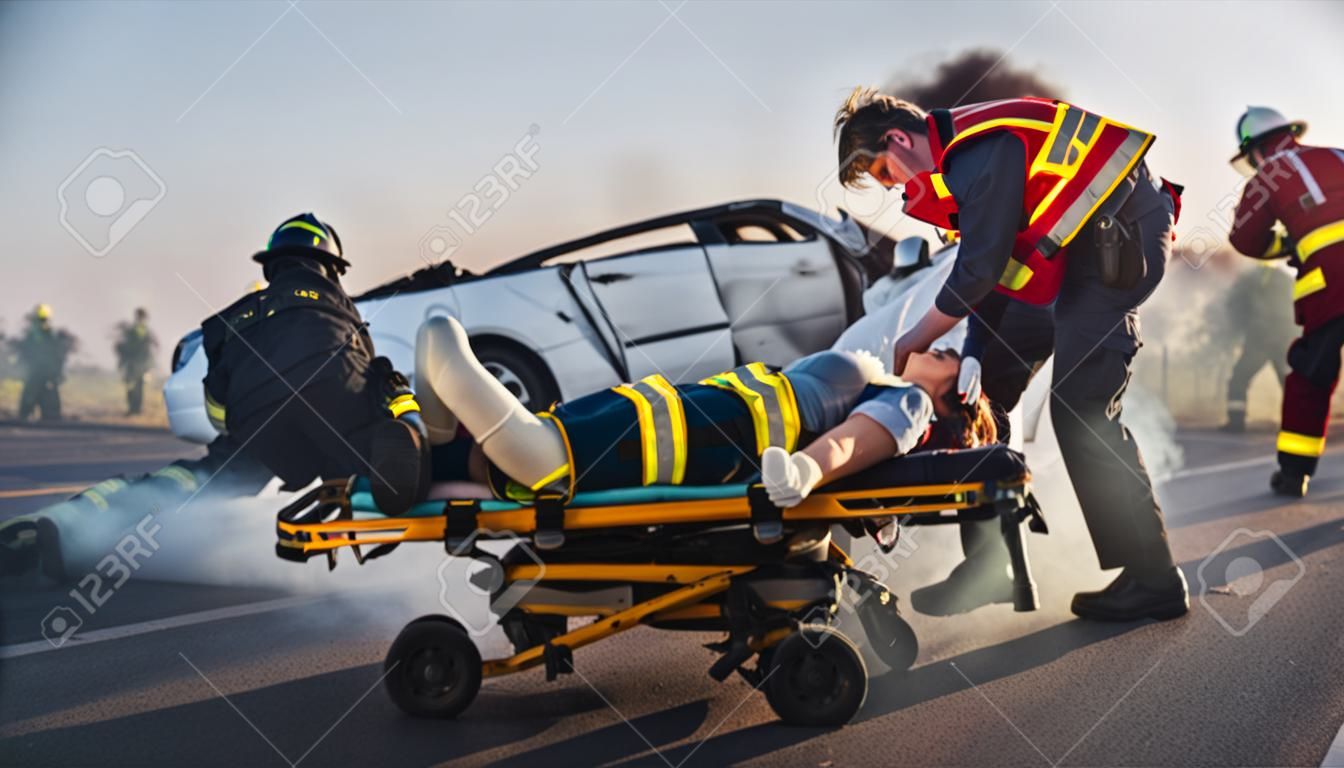 On the Car Crash Traffic Accident Scene: Paramedics Save Life of a Female Victim Lying on Stretchers. They Listen To a Heartbeat, Apply Oxygen Mask and Give First Aid. Firefighters Extinguish Fire