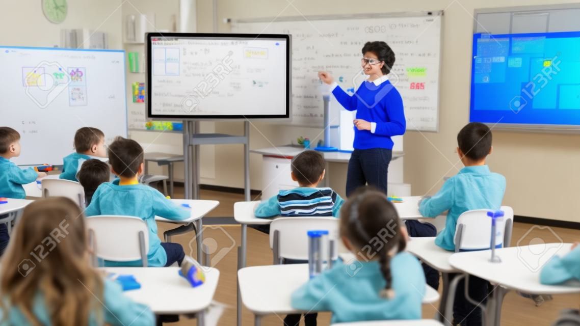 O professor de ciências da escola primária usa o quadro branco digital interativo para mostrar à sala de aula cheia de crianças como a programação de software funciona para a robótica.