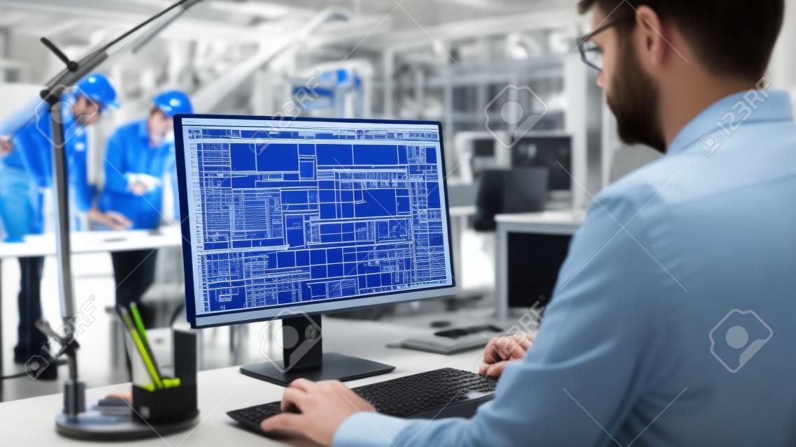 Over de schouder Shot van Engineer Werken met CAD Software op Desktop Computer, scherm toont technische ontwerpen en tekeningen. In de achtergrond Engineering Facility gespecialiseerd in industrieel ontwerp