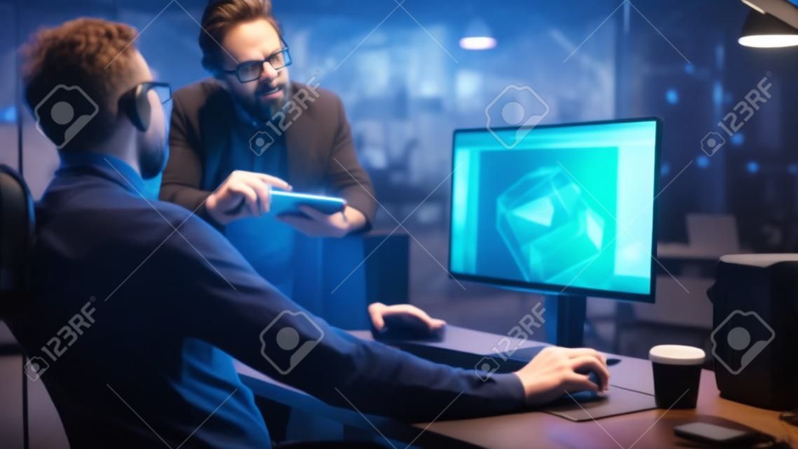Wieczorem kreatywny twórca gier komputerowych w średnim wieku pracuje na komputerze stacjonarnym z ekranem projektowania poziomów gier wideo 3D, rozmawia z młodszym specjalistą, który korzysta z komputera typu tablet
