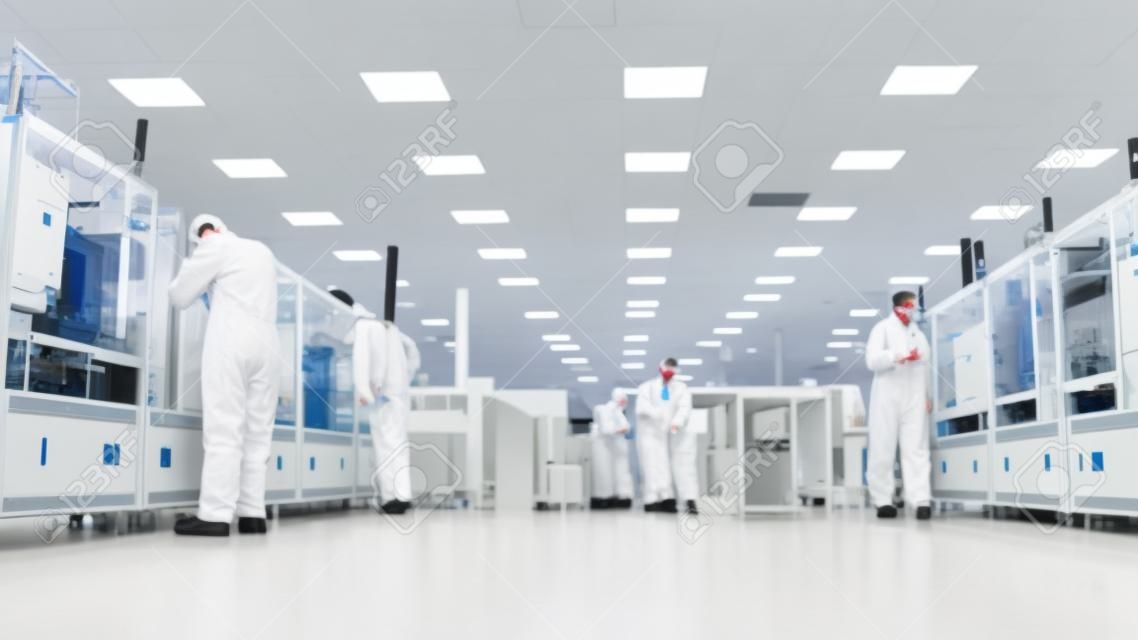 Zespół naukowców w sterylnych kombinezonach pracujących z komputerami, mikroskopami i nowoczesnymi maszynami przemysłowymi w laboratorium. proces wytwarzania produktu: farmaceutyki, półprzewodniki