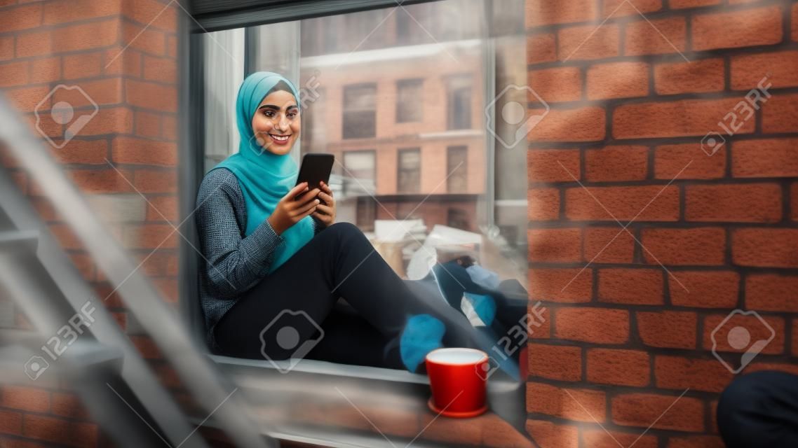 Portret młodej muzułmanki korzystającej ze smartfona, siedzącej na parapecie okna w przytulnym mieszkaniu z piaskowca. dziewczyna sprawdza media społecznościowe online. kąt kamery od