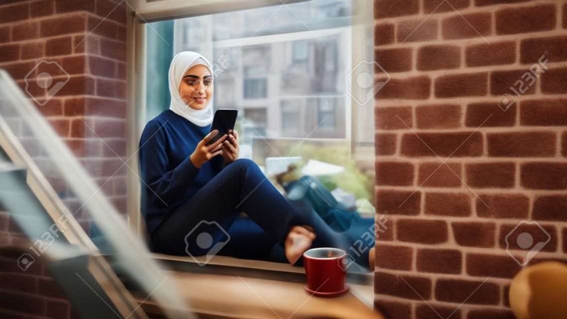 Porträt einer jungen muslimischen frau, die ein smartphone benutzt, während sie auf einer fensterbank in einer gemütlichen brownstone-hauswohnung sitzt. mädchen überprüft online-social-media. kamerawinkel von